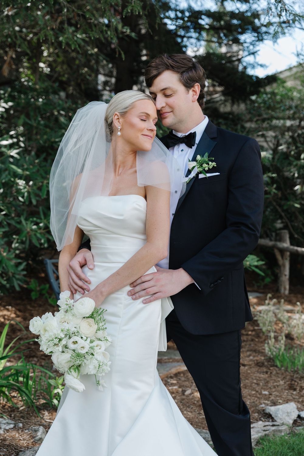Newlywed couple shares intimate moment outdoors with bride in white gown holding white bouquet and groom in black tuxedo.