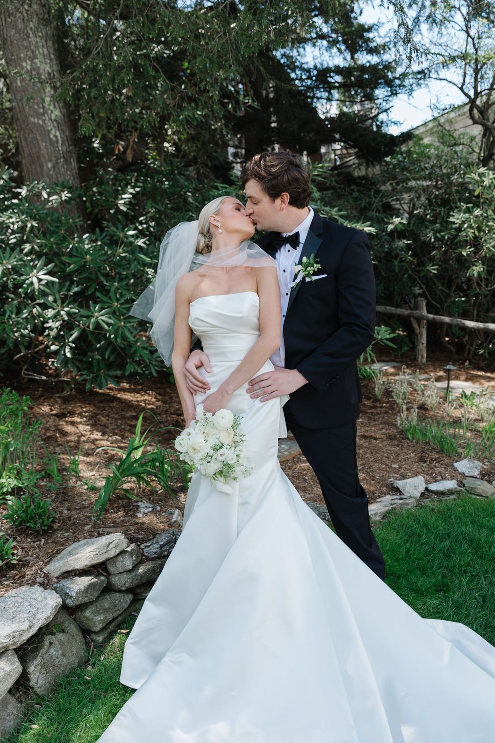 A romantic wedding portrait showing a bride in white gown and groom in black suit embracing in a lush garden setting.