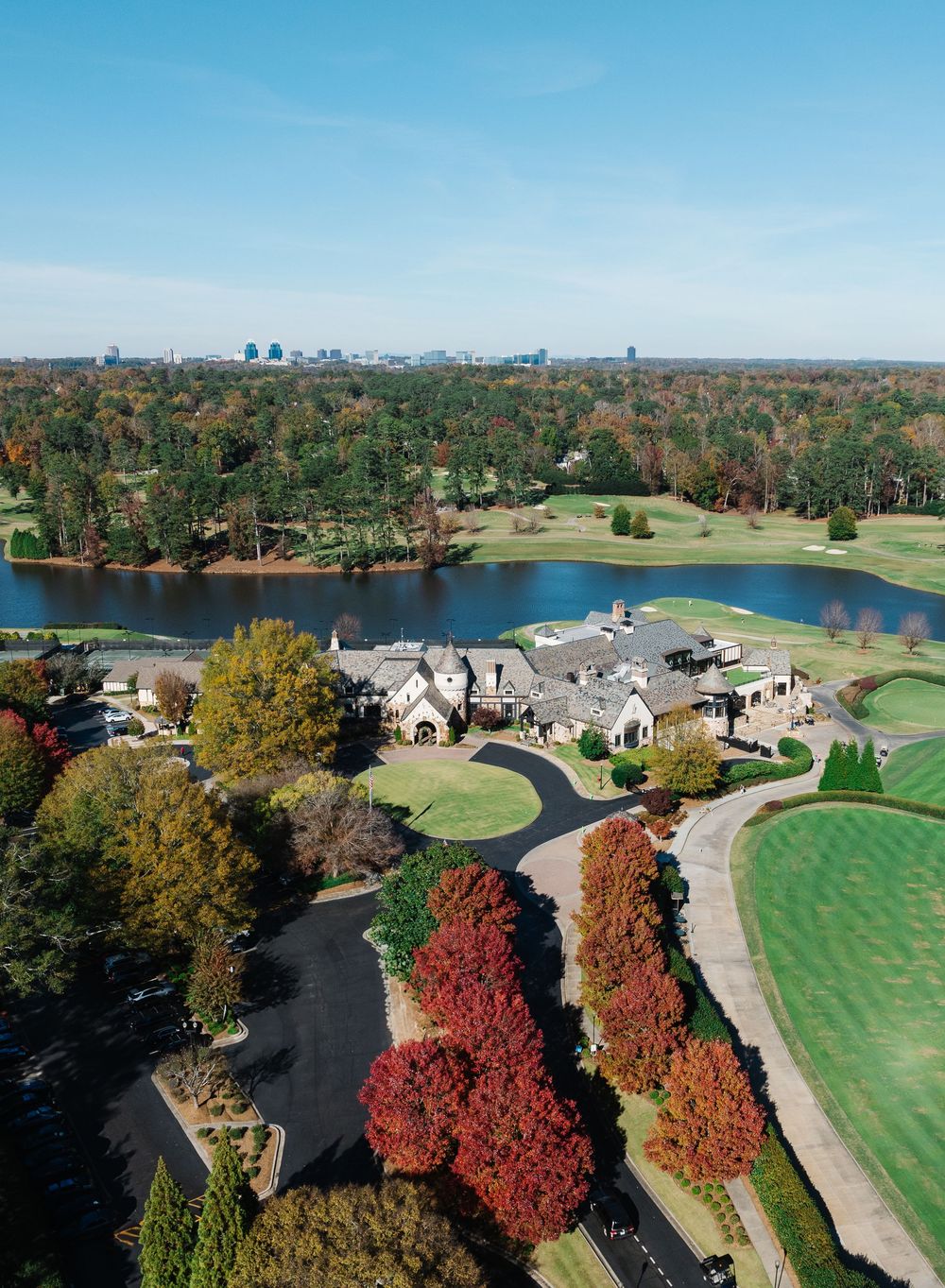 Aerial view of a luxury golf course clubhouse surrounded by autumn trees, green fairways and a scenic lake on a clear day.