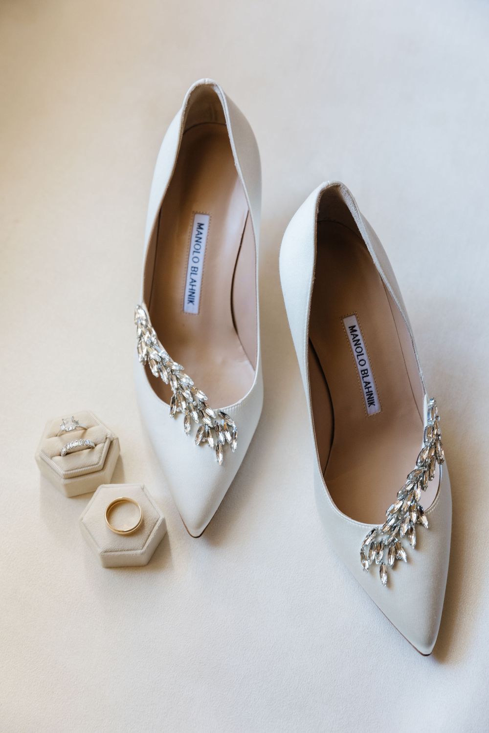 Elegant white satin wedding heels with crystal embellishments sit beside small ring boxes on a light background.