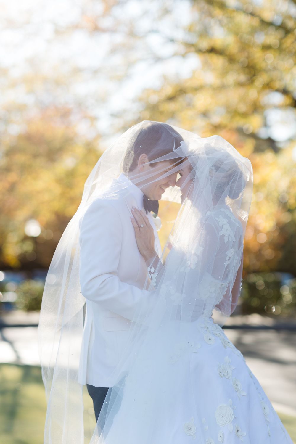 A romantic wedding couple share a tender moment outdoors with sunlit autumn foliage in the background.