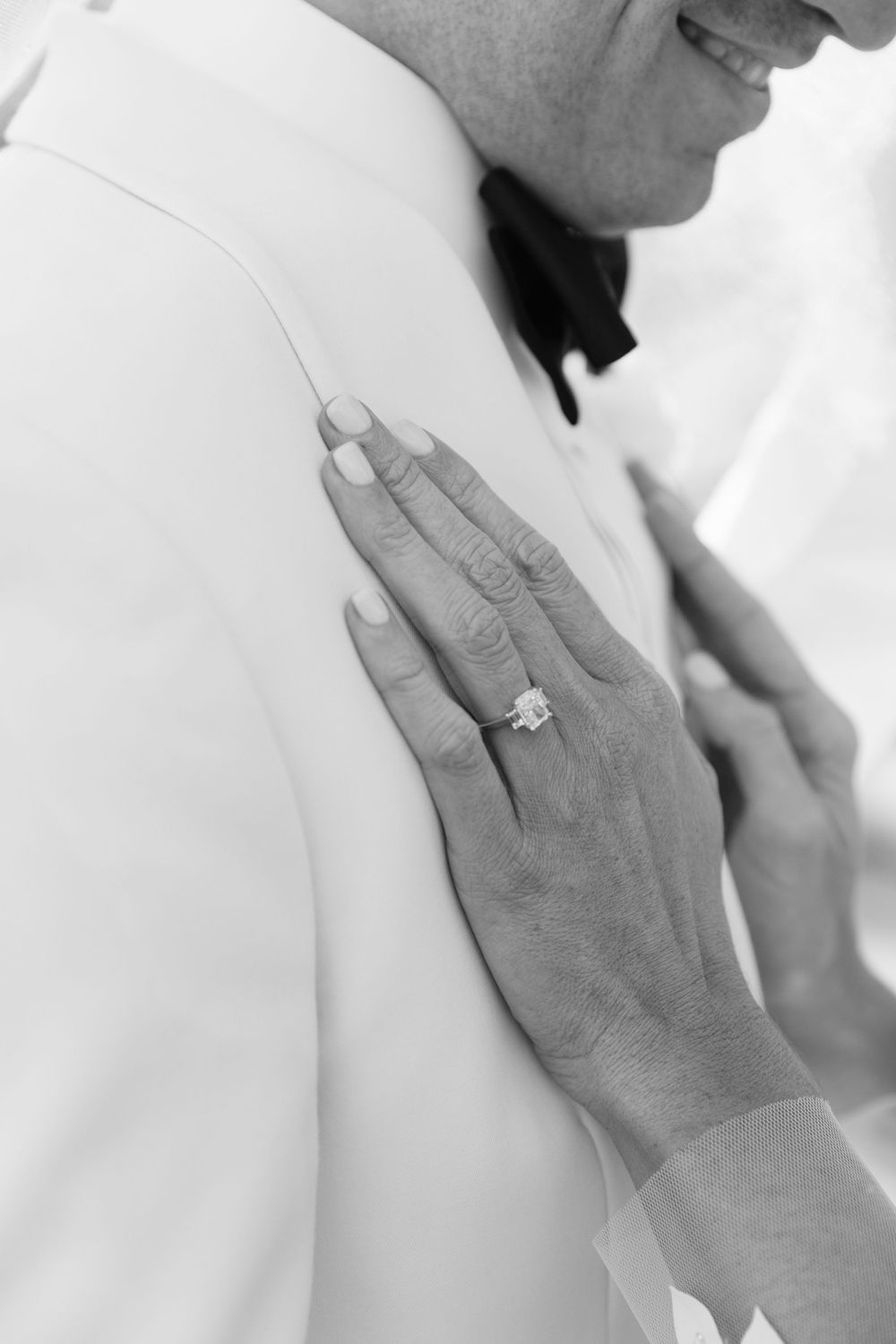 Black and white photo of a hand with an engagement ring resting on someone's shoulder showing tender affection.