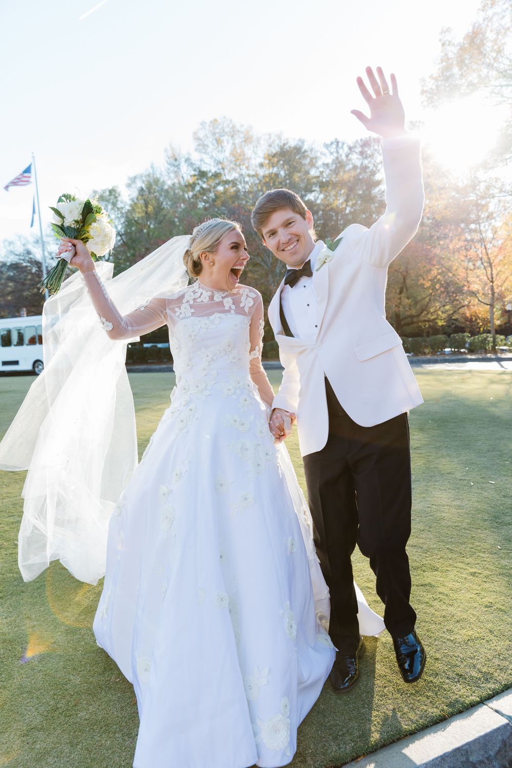 A joyful newlywed couple celebrating outside on their wedding day with a long white gown and classic white tuxedo jacket.
