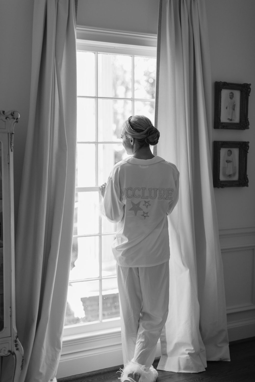 A figure in white loungewear gazes through a tall window with elegant curtains in a black and white composition.