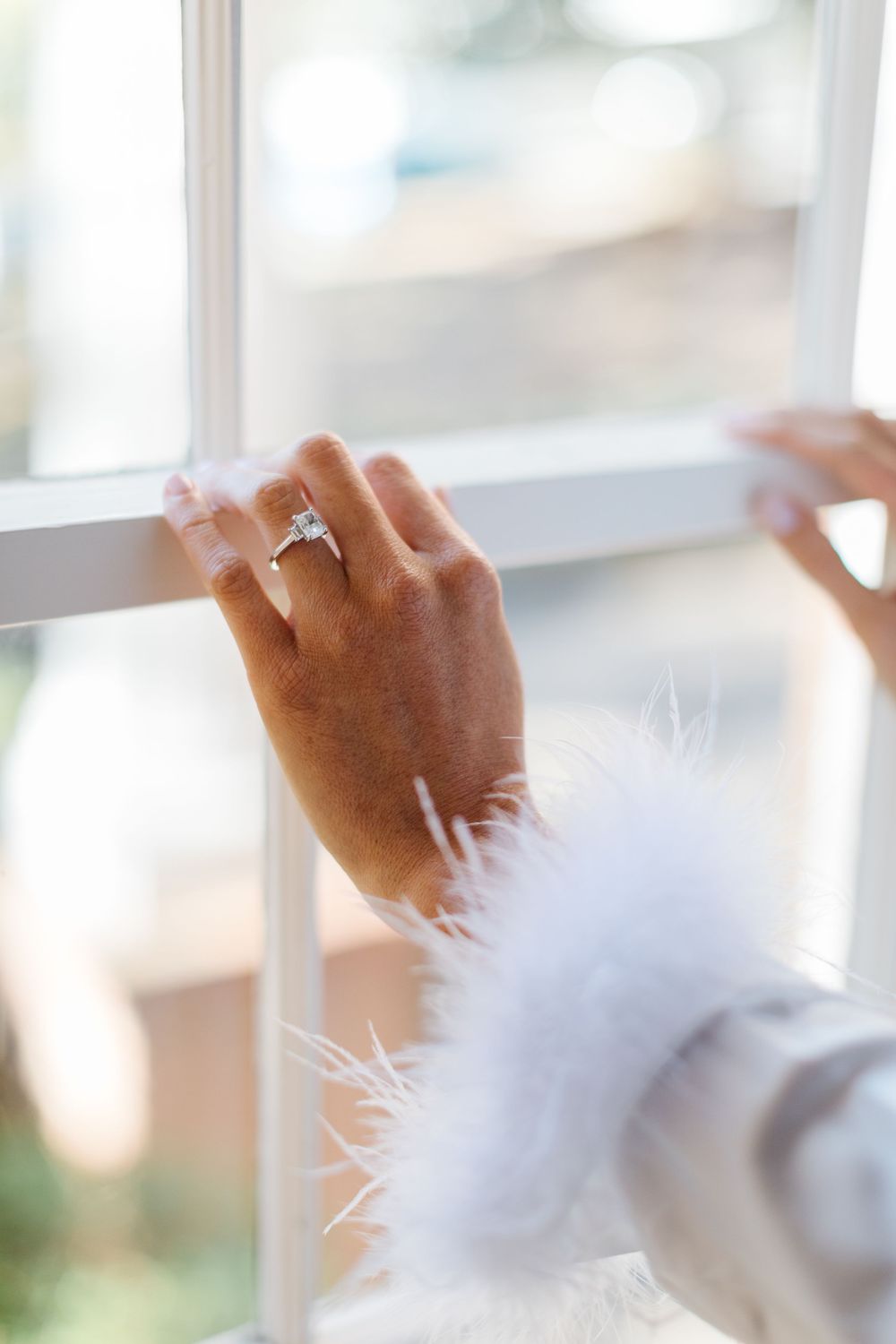 A hand wearing an engagement ring gently touches a bright window frame, with sunlight streaming through the glass.