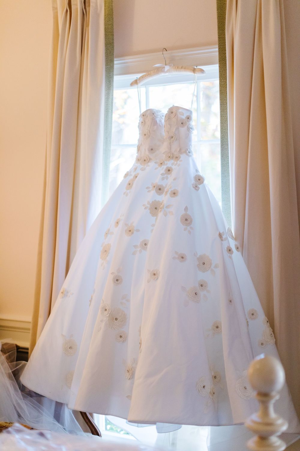 A white wedding gown with floral embellishments hangs in a window between cream colored curtains.