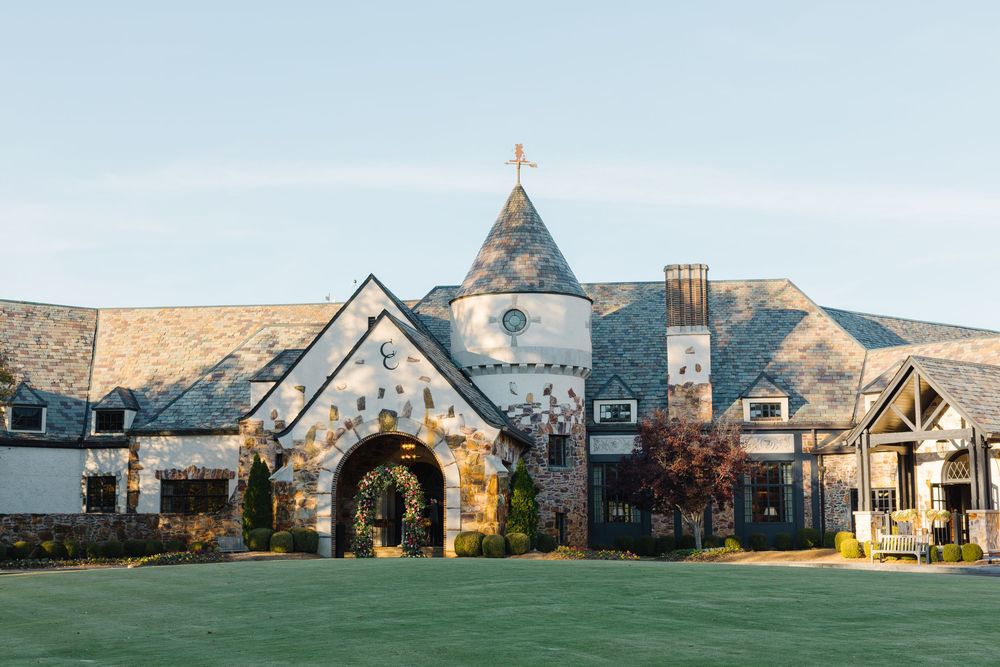 A grand stone capital city country club with an arched entrance and cross-topped steeple stands against a light blue sky.