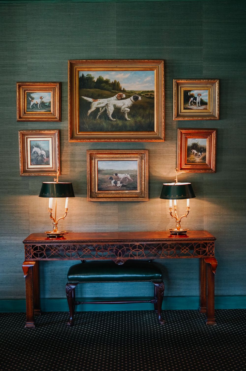 Elegant console table with antique dog portrait paintings and brass candelabra lamps against teal textured wallpaper.