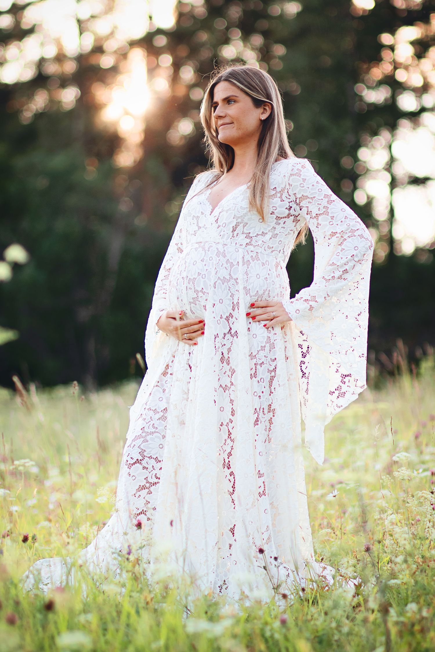 Pregnant woman in white lace bell-sleeve gown standing in sunlit wildflower meadow.