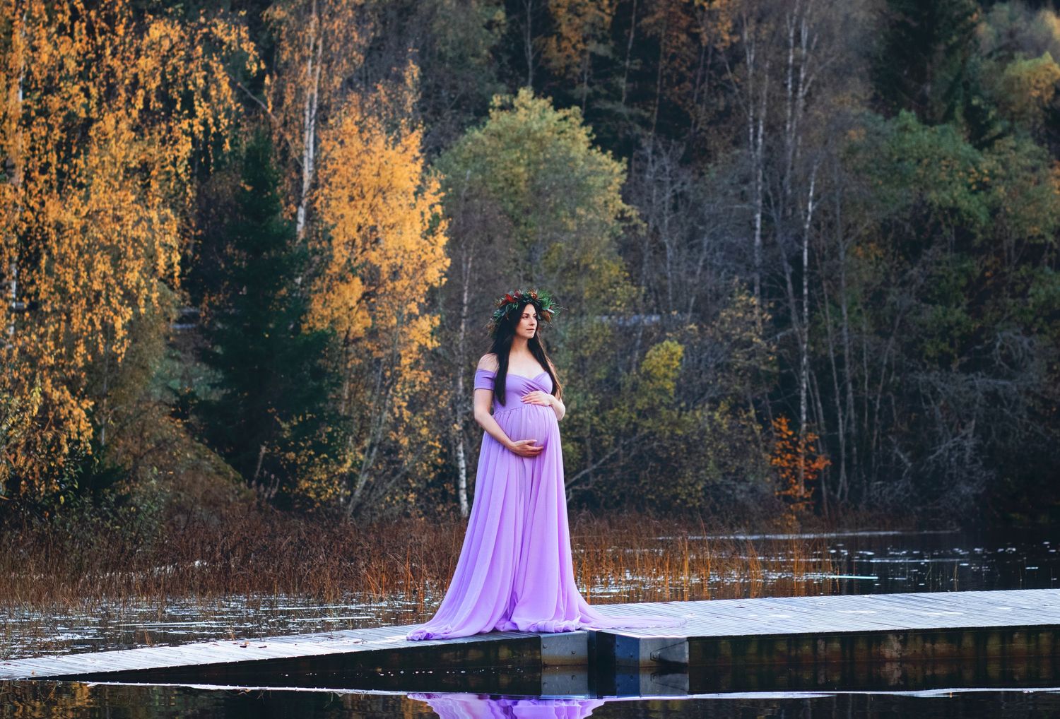 Pregnant woman in lavender gown standing on dock surrounded by autumn foliage.