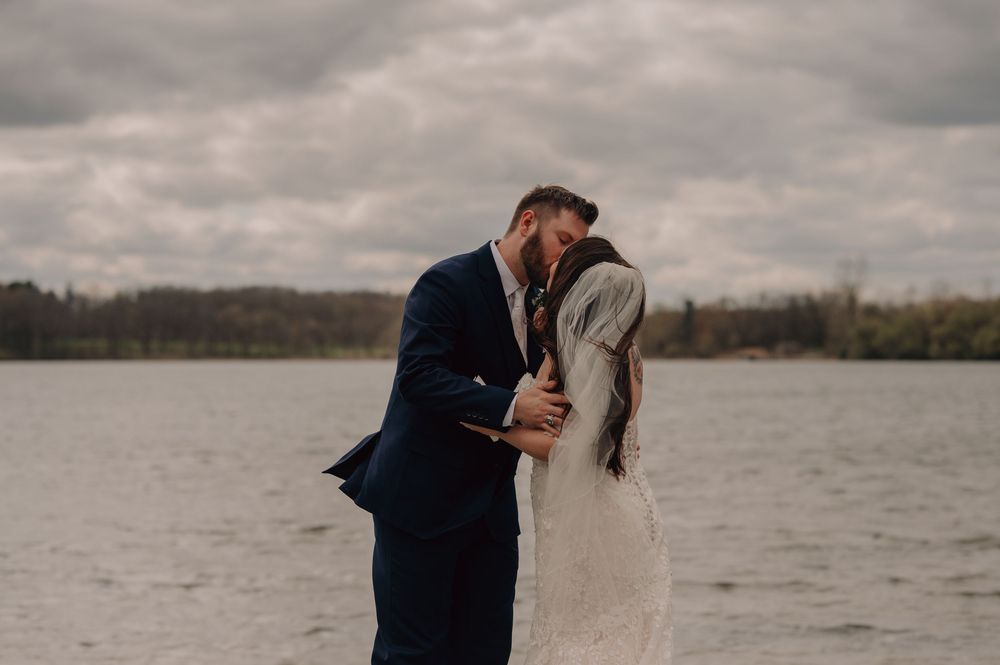 A couple shares a romantic kiss on a beach with stormy clouds and a lake in the background on their wedding day.