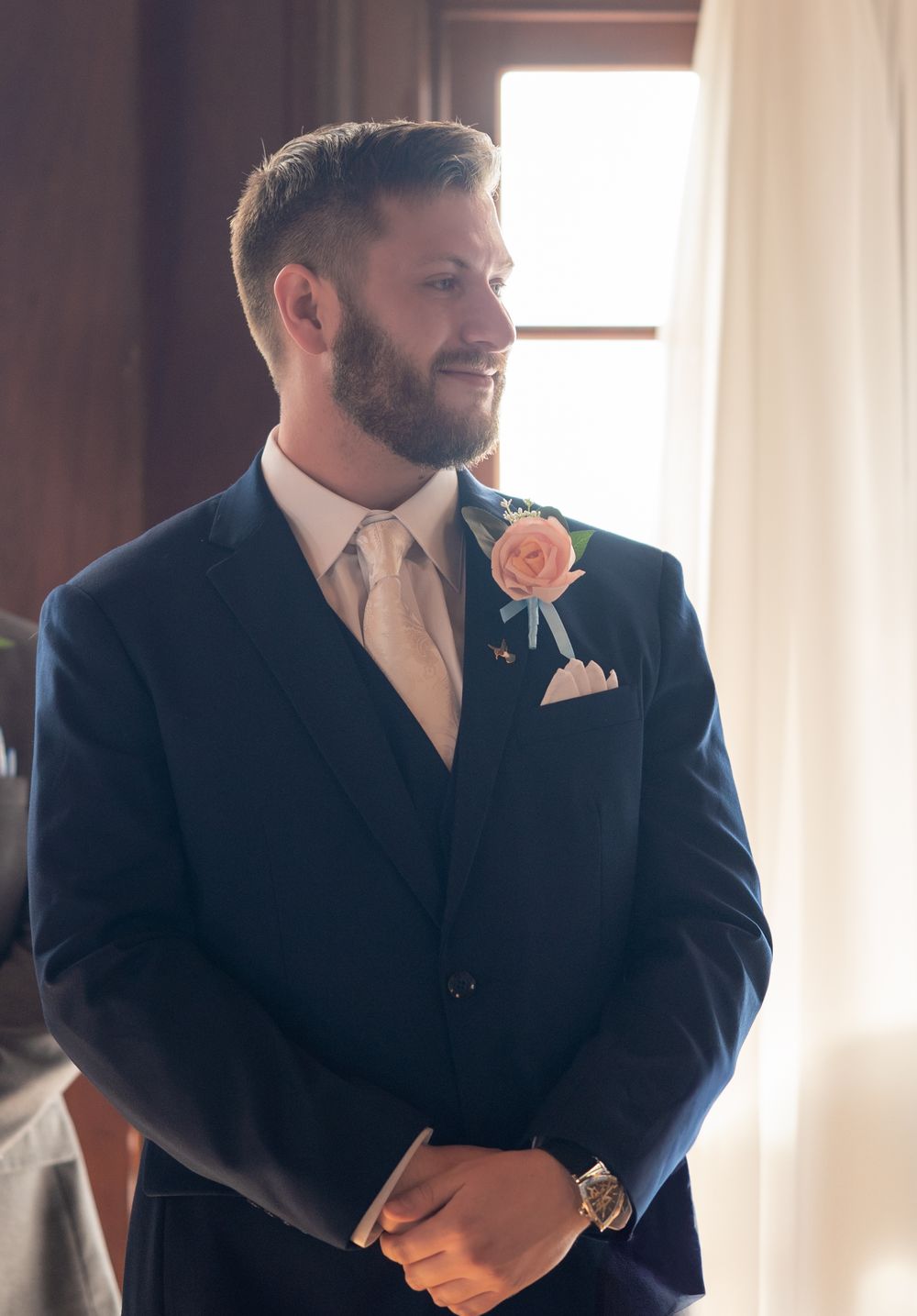 Well-dressed groomsman in navy suit with peach boutonniere smiles while standing near window at formal event.