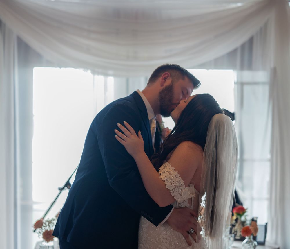 A tender moment between newlyweds sharing their first kiss under elegant white drapery at their wedding ceremony.
