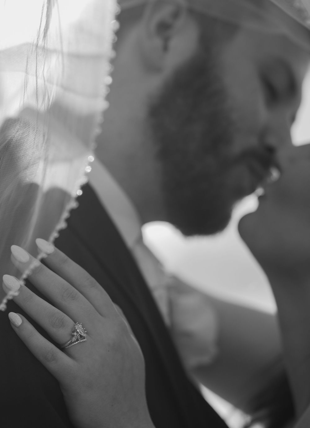 A dramatic black and white portrait showcases a delicate veil and engagement ring during an intimate wedding moment.