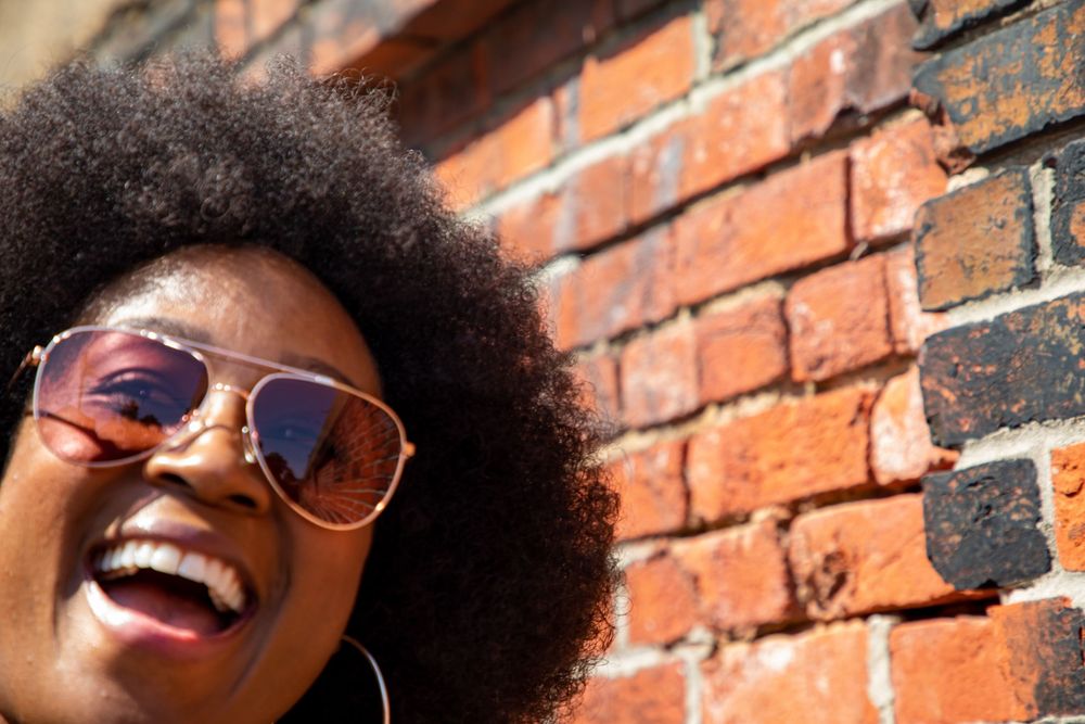 A joyful expression and natural afro hairstyle against a rustic brick wall background, wearing stylish sunglasses.