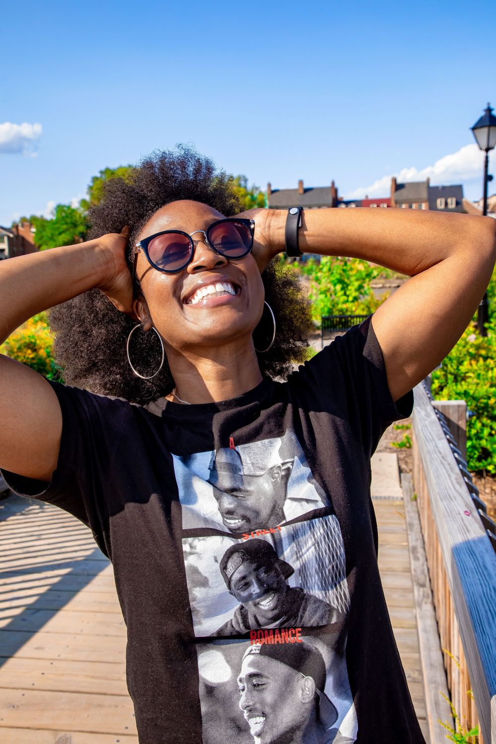 Person in sunglasses and a graphic t-shirt poses on a bridge with natural scenery in the background on a sunny day.