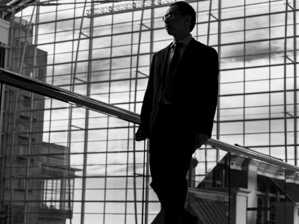 Black and white silhouette of a businessman standing on modern office stairs against geometric window backdrop.