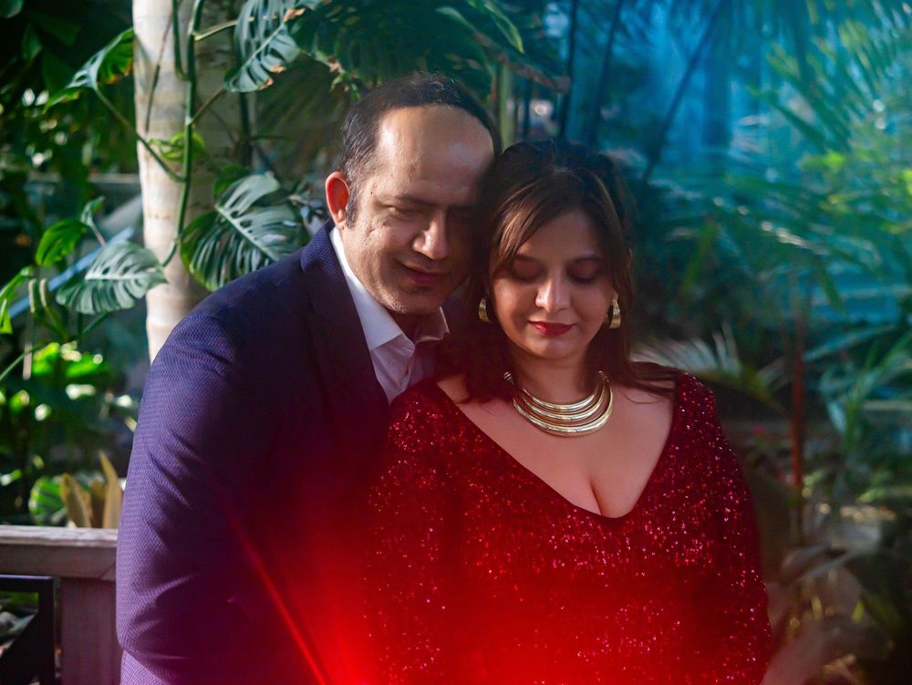 A couple poses for a formal portrait, with the woman wearing a red sparkly dress and pearl necklace against a tropical backdrop.