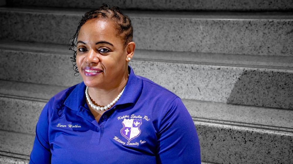 Athletic staff member in royal blue polo shirt with embroidered logo smiles while seated on concrete steps.