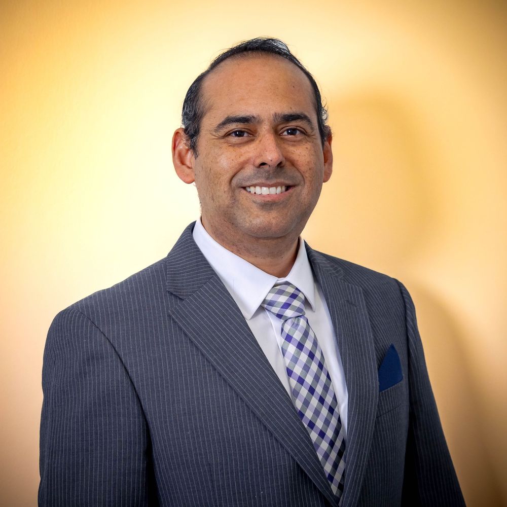 Professional headshot of person in gray pinstripe suit with purple checkered tie against warm gold background.