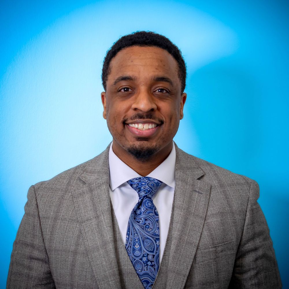 Professional portrait of someone in a grey plaid suit and blue patterned tie against a bright blue background.