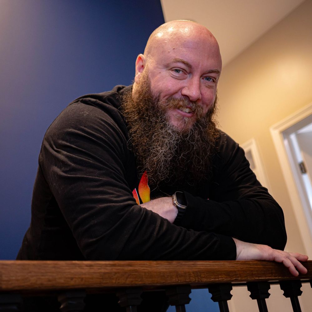 Cheerful bearded man in black shirt leaning on wooden railing in indoor setting.
