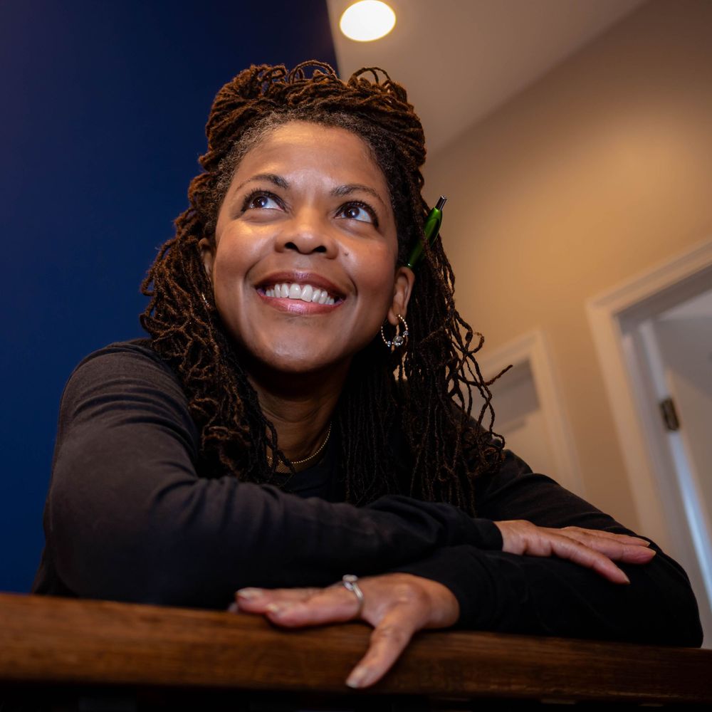 Smiling person in a black sweater leaning on wooden railing with a thoughtful expression in an indoor setting.