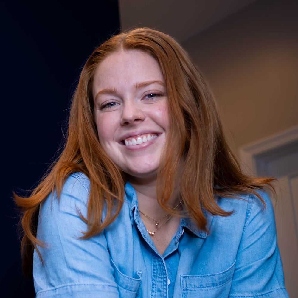 Young woman with flowing red hair wearing a light blue denim shirt smiles warmly at the camera.