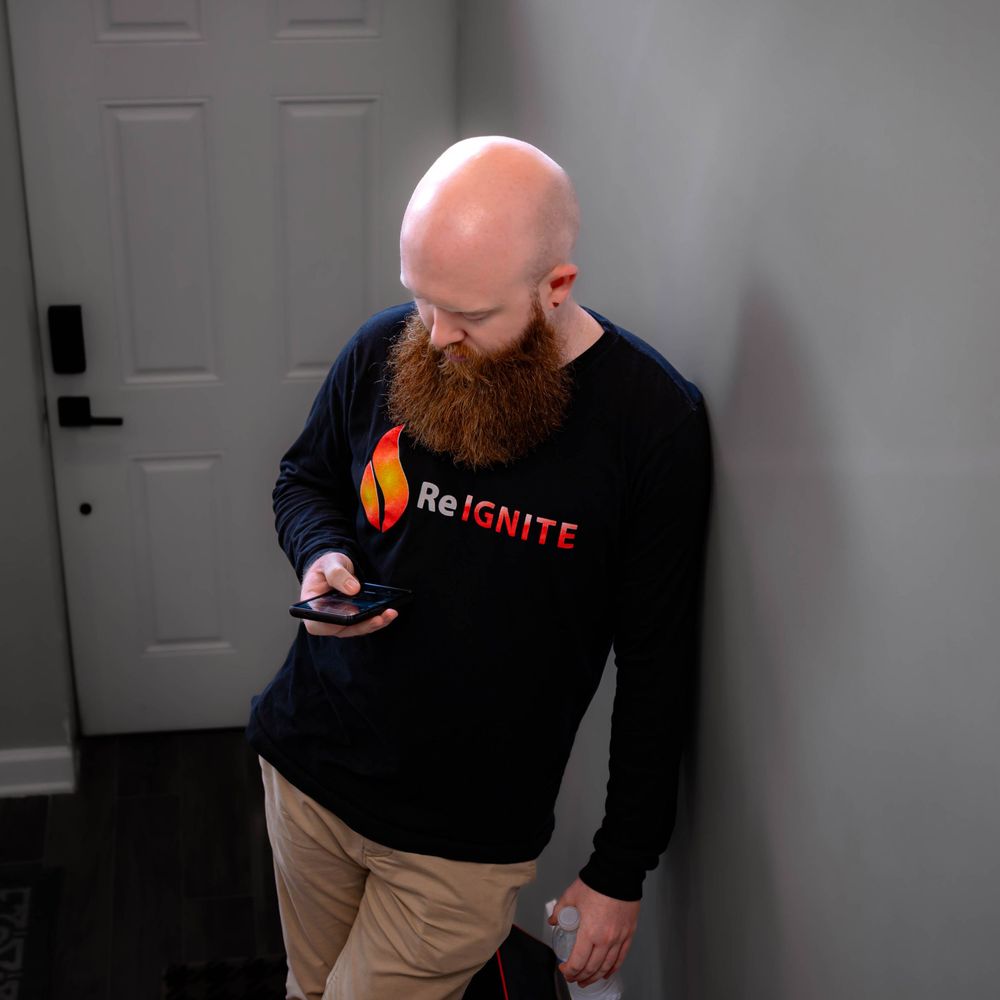 A person in a black ReIgnite sweater checking their phone while standing against a white door.