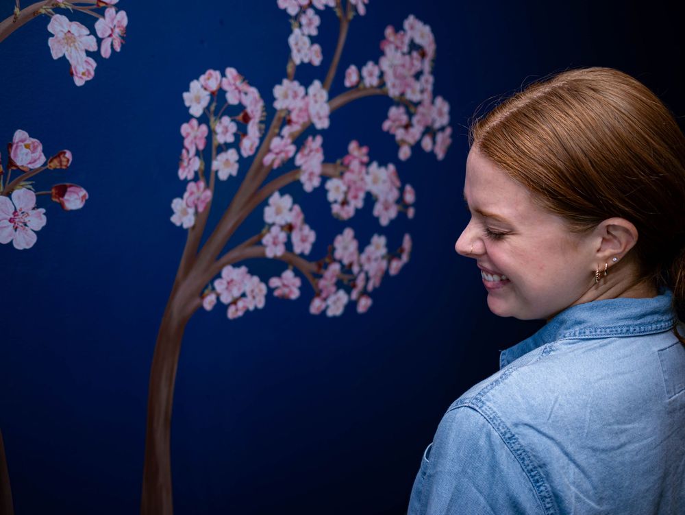 A person in denim shirt smiles while standing next to a painted cherry blossom tree mural against dark blue wall.