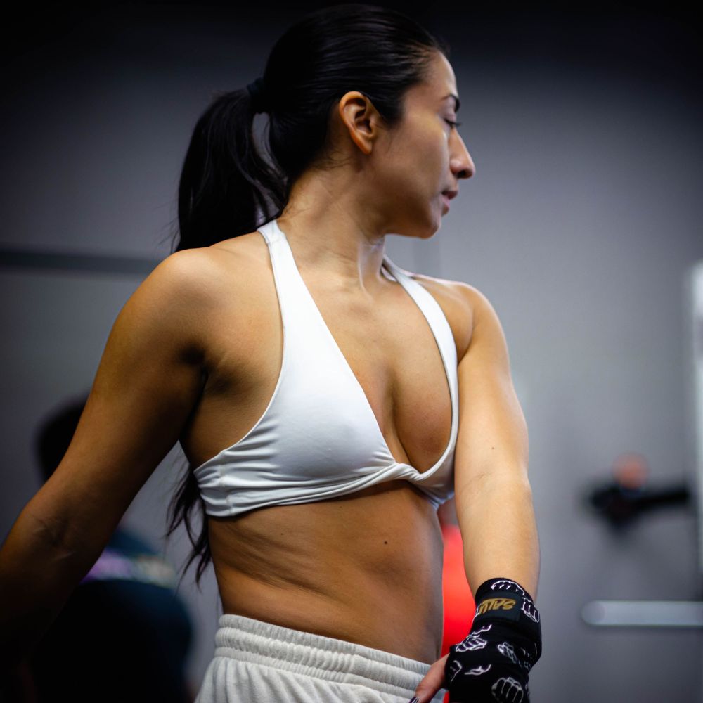 Side view of an athlete in white sports bra and gloves taking a break during a workout in a dark gym setting.
