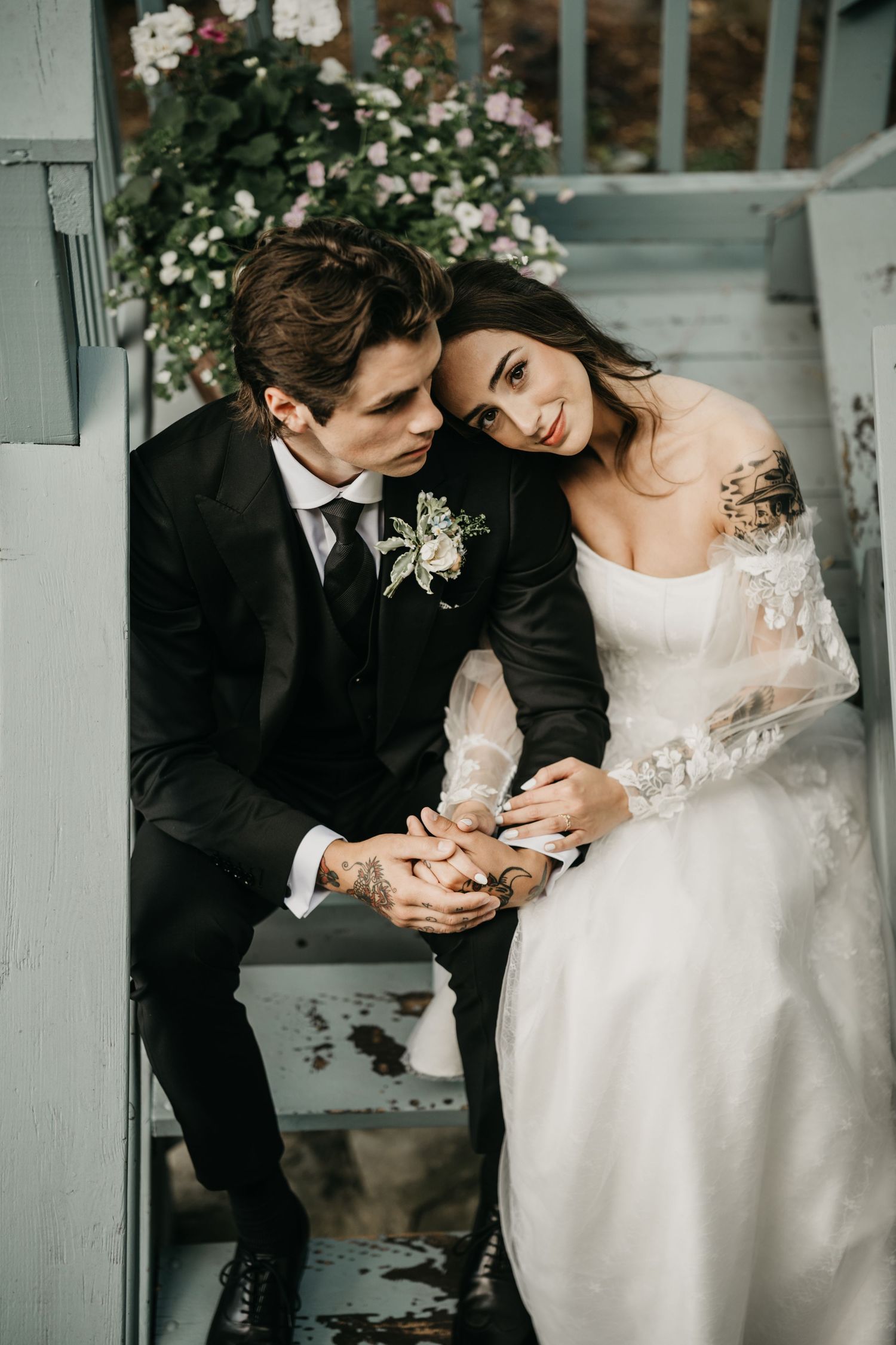 A couple in wedding attire sits intimately on light blue painted wooden stairs beside a tree.
