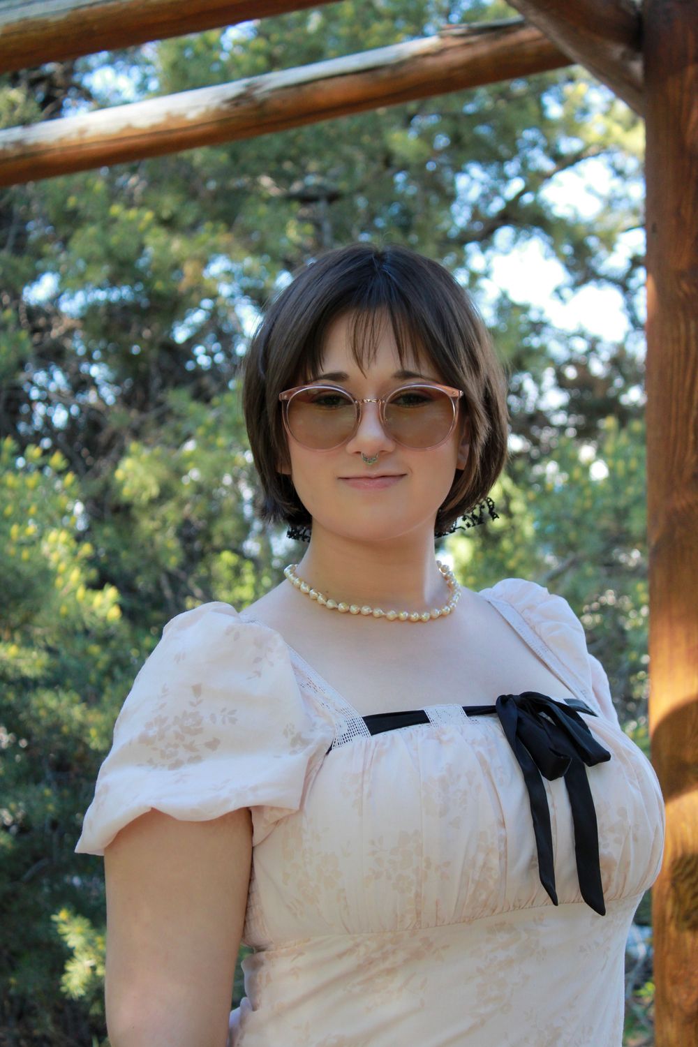 Person in a white vintage-style dress with black bow detail wearing sunglasses and pearl necklace outdoors under wooden pergola.