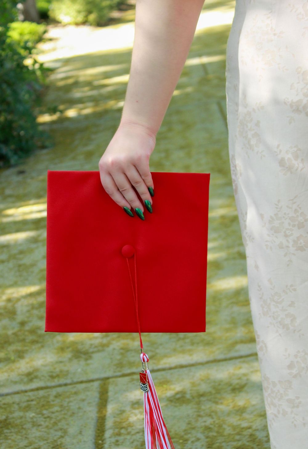 A hand with dark nail polish holds a bright red graduation cap with tassel against a blurred outdoor background.