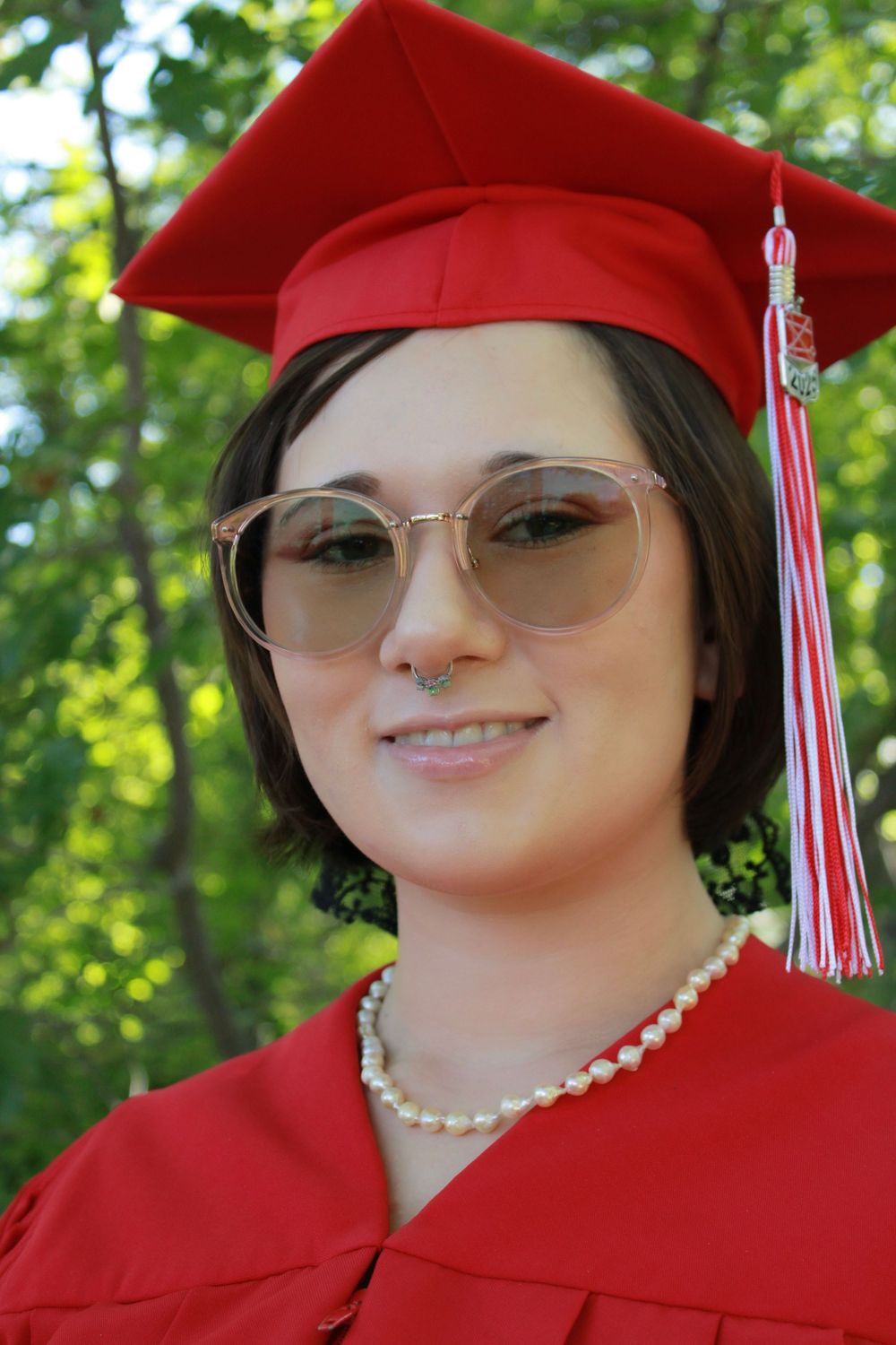 Graduate in red cap and gown wearing glasses and pearl necklace poses for portrait against green outdoor background.