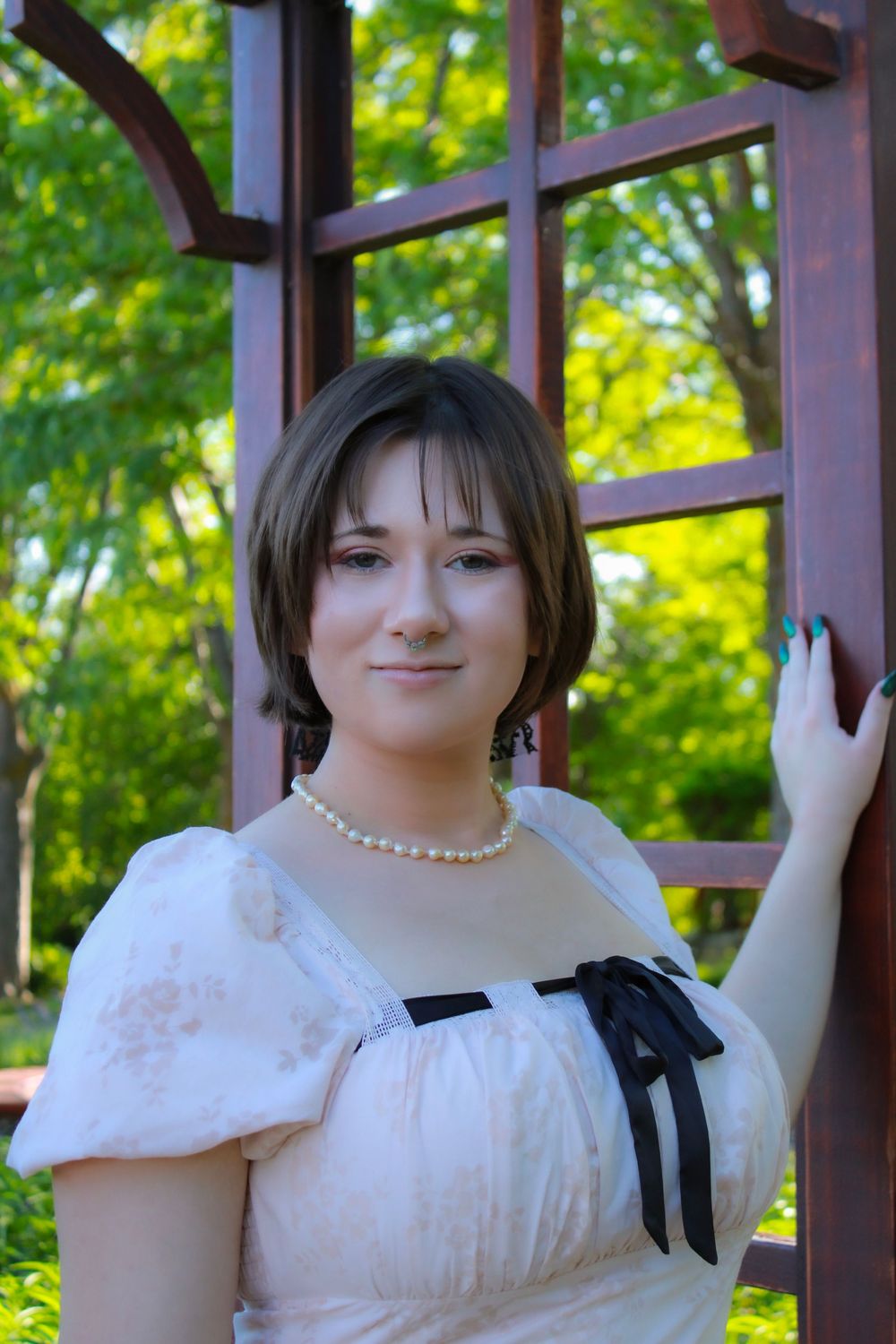 A person in a white Regency-style dress with a pearl necklace and black bow stands in front of a garden trellis.
