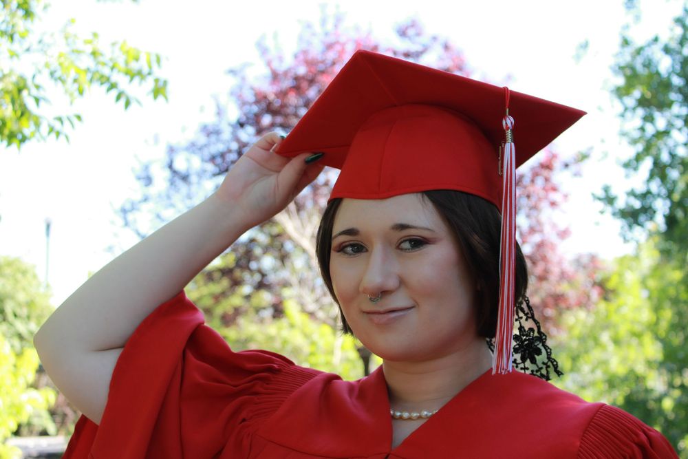 Graduate in bright red cap and gown adjusts their mortarboard while posing outdoors on a sunny day with trees in background.