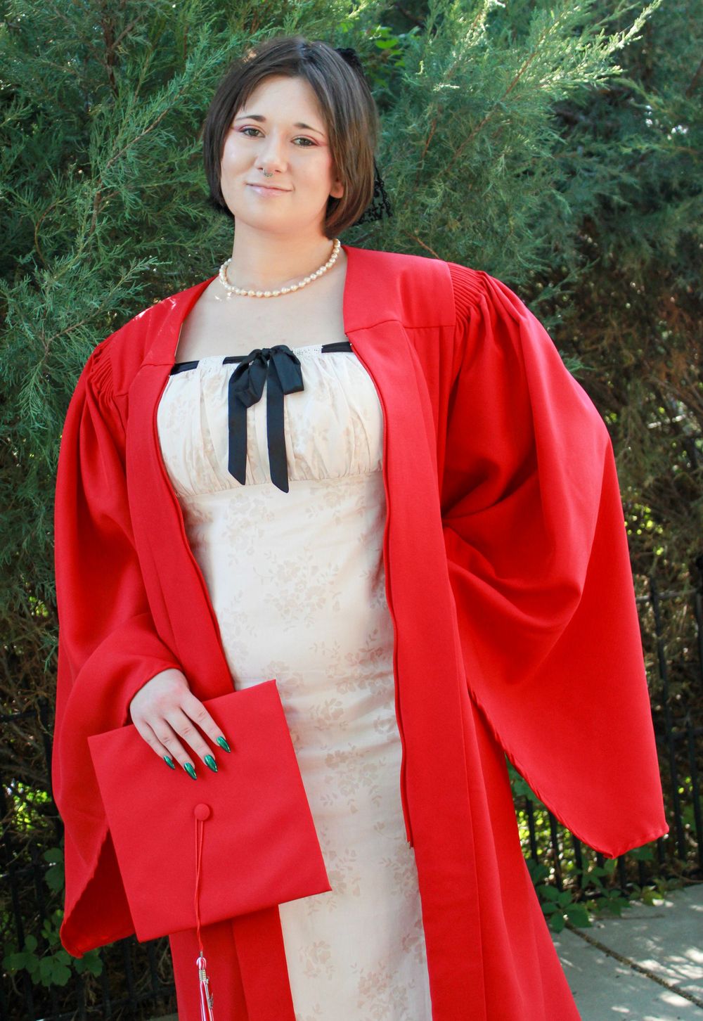 A graduate poses in a bright red academic gown and cream-colored dress with a black bow tie detail while holding a red cap.