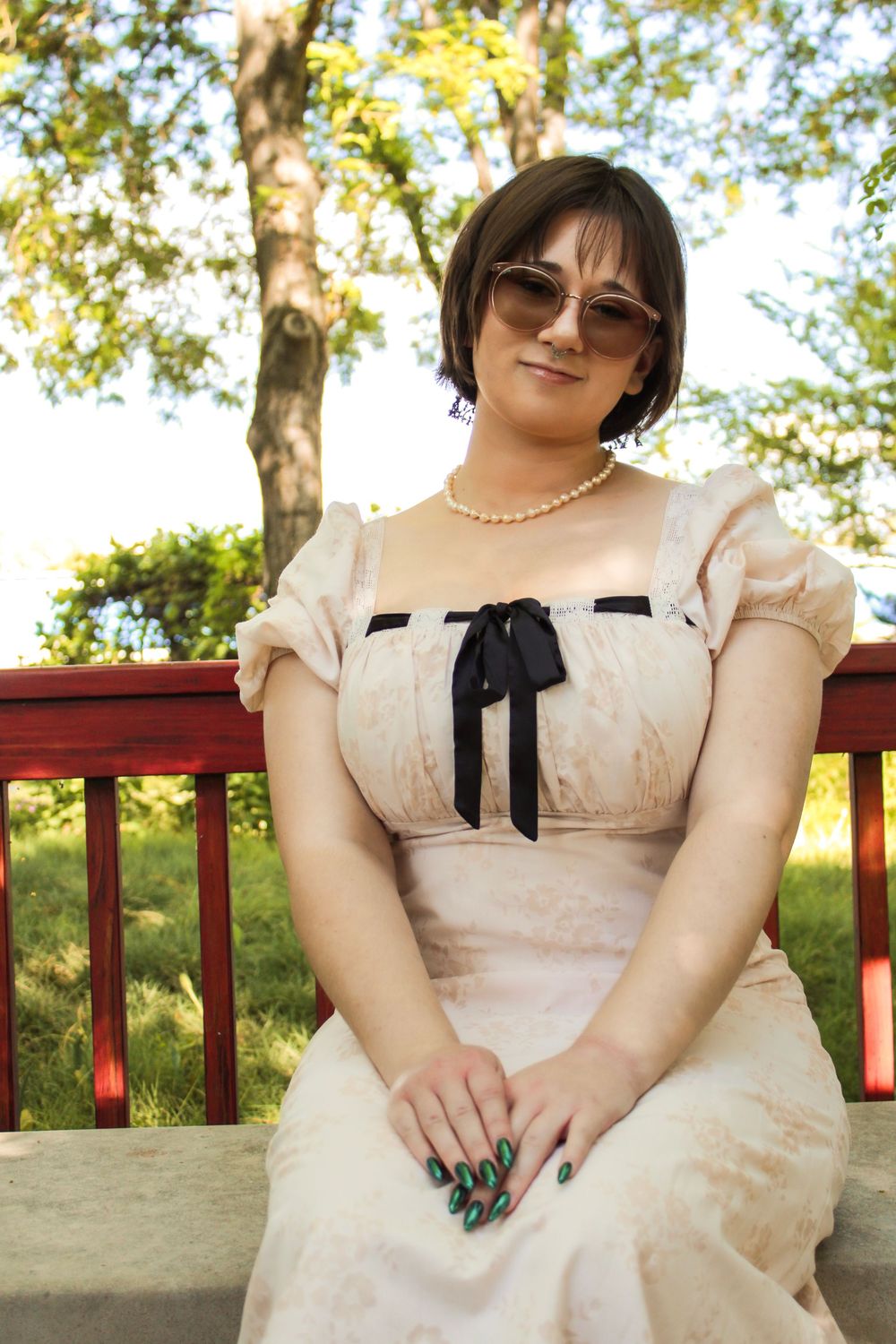 A vintage-inspired cream dress with black bow accent is worn with sunglasses and a pearl necklace while seated outdoors.