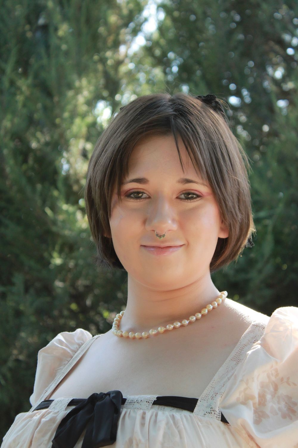 A person wearing a white dress with black ribbon trim and pearl necklace posed outdoors against greenery.