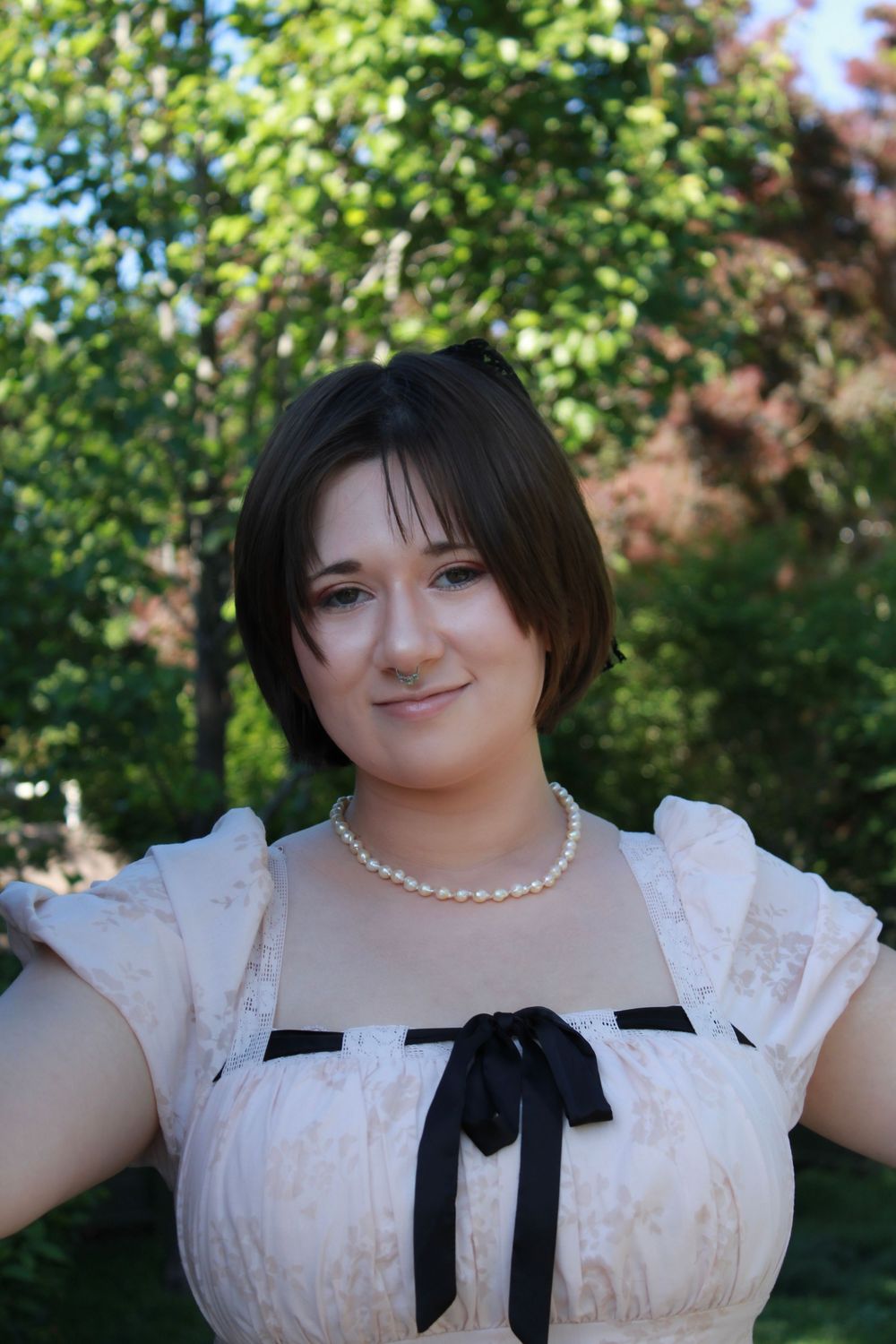 Person wearing a white vintage-style dress with black bow and pearl necklace stands outdoors in a garden setting.