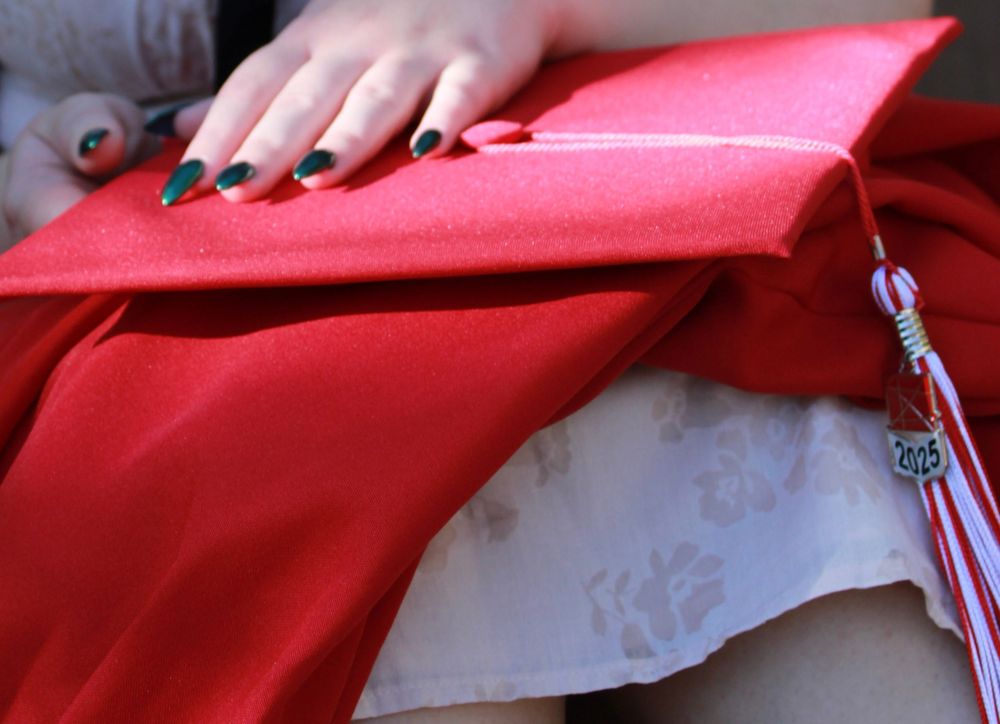 Black manicured hands rest on a red graduation cap and gown with a tassel visible in the corner.