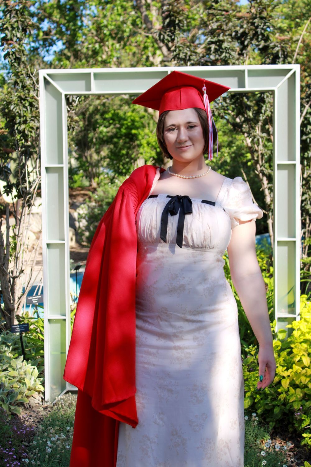 A graduate in a red cap and gown poses outdoors in a white dress under a decorative garden arch.
