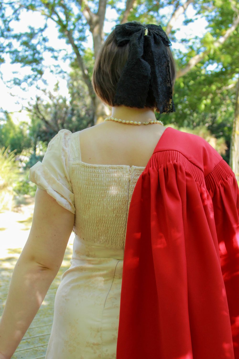 Back view of someone wearing a cream colored dress with pearls and carrying a bright red draped garment outdoors.