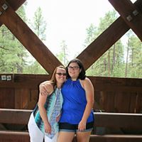 Two women smiling together under rustic wooden bridge structure outdoors.