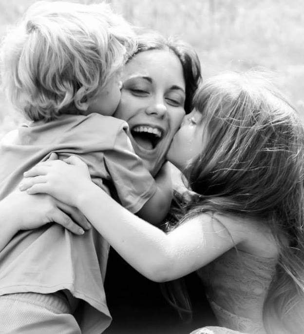 Black and white photo of two children kissing and hugging a laughing mother outdoors.
