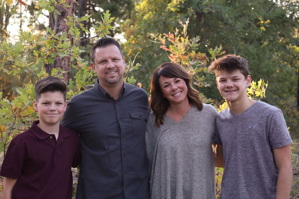 Family of four smiling together outdoors among colorful autumn foliage trees.