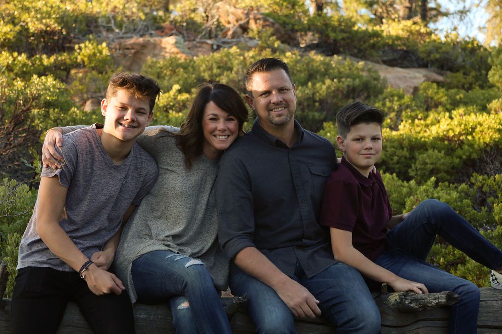 Happy family of four smiling outdoors in a sunny natural setting with green shrubs.