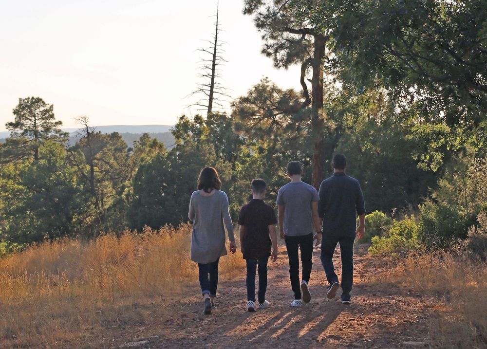 Family of four walking together on a dirt trail through a scenic forested landscape.