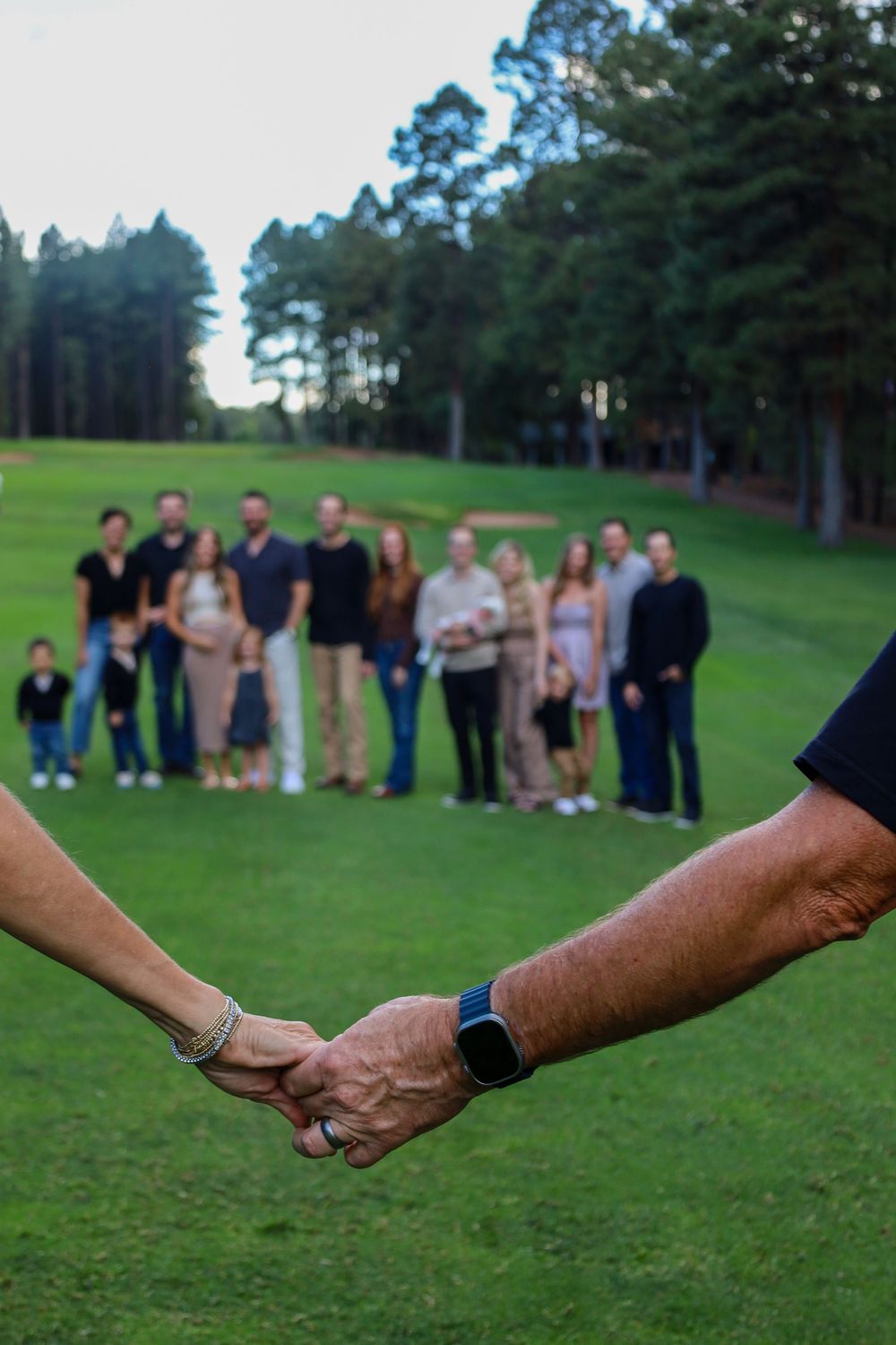 Close-up of older couple holding hands with large extended family group visible in background on lawn.